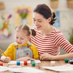 A woman wit a red and white striped top sits with a young child. They smile and draw and play at a table.