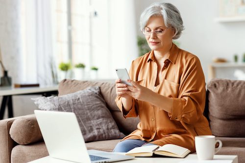 A middle aged woman sits at home on her phone and computer studying